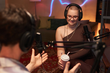 Medium full shot of smiling young girl podcaster listening to male guest answer while sitting in...
