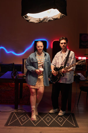 Full shot of young girl holding plastic cup and adult male with semi-acoustic guitar standing in studio with neon light and looking at cameraの写真素材