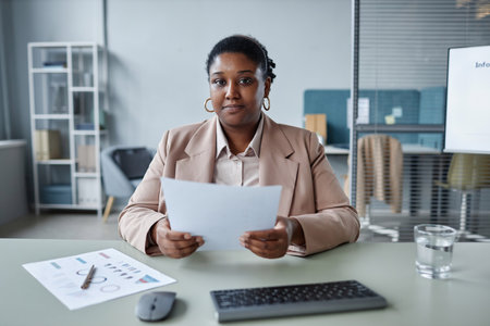 Portrait of adult African American businesswoman wearing pastel suit jacket holding paper while sitting at office desk and looking at cameraの写真素材