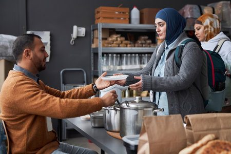 Side view portrait of Middle Eastern woman wearing hijab receiving hot meal at soup kitchen or...