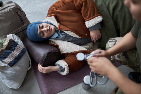 High angle portrait of adult Middle Eastern woman wearing headscarf and talking to doctor at refugee shelter while lying on floorの写真素材
