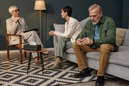 Full length portrait of senior man holding glass of water sitting on couch with wife during couples therapy session or counselling, copy spaceの写真素材