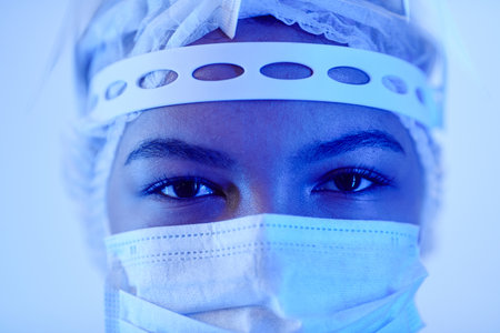 Extreme close up shot of dark-eyed female doctor n blue neon light wearing medical mask and mop cap...
