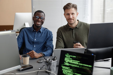 Portrait of cheerful African American IT specialist in glasses and Caucasian male coworker holding takeaway coffee cup looking at camera while sitting at deskの写真素材