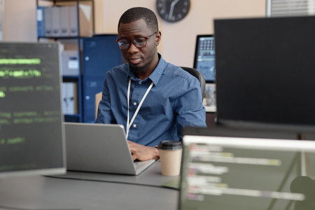 Medium shot of focused African American male programmer in glasses typing on laptop while sitting at...