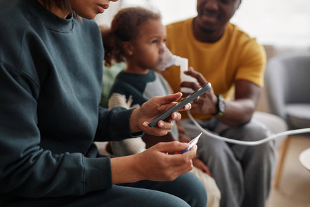 Side view of unrecognizable mom holding smartphone and thermometer while dad and daughter sitting in...