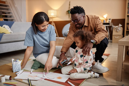 Full shot of smiling African American mother and father pointing at paper on floor and asking little daughter holding marker about her drawingの写真素材