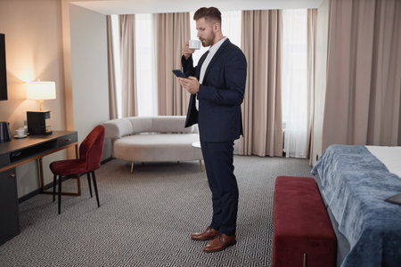 Full length portrait of elegant young businessman wearing suit and enjoying cup of coffee in hotel room during business travel, copy spaceの写真素材