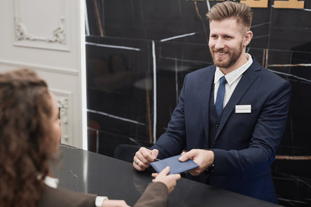 Portrait of smiling young man taking documents of hotel guest for check in and register at reception deskの写真素材