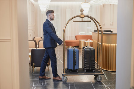 Side view portrait of male porter pushing cart with with bags and luggage in luxury hotel