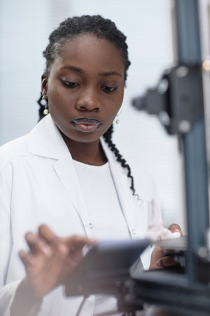 Medium close up shot of young Black woman technician working with 3D printer in laboratoryの写真素材