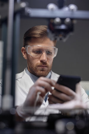 Medium close up shot of Caucasian male engineer wearing safety glasses looking at defocused 3D printer controller in handsの写真素材