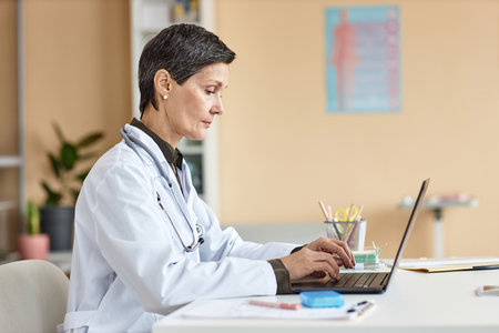 Side view of focused senior Caucasian woman doctor typing on laptop while sitting in clinic office