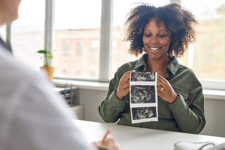 Medium shot of cheerful pregnant Black woman looking at unborn baby sonogram image during consultation in prenatal clinicの写真素材