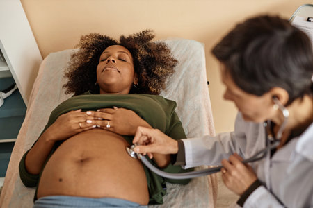 Top down view of pregnant African American woman lying on clinic bed with closed eyes while unrecognizable female obstetrician conducting abdomen auscultationの写真素材