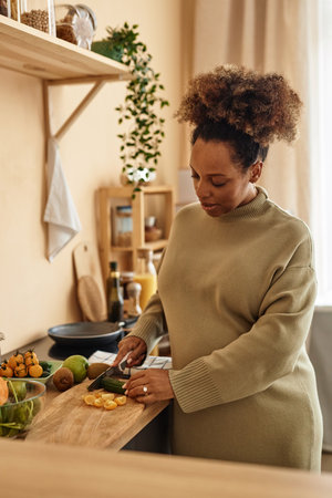 Vertical shot of pregnant Black woman wearing pastel green sweater dress cutting vegetables for salad on wooden board in kitchenの写真素材