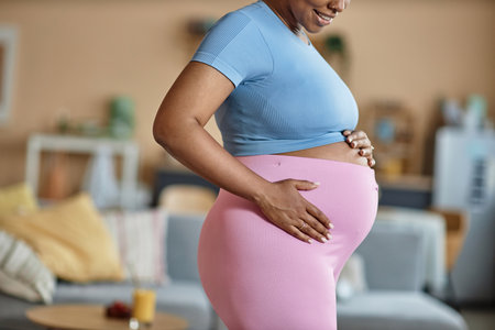 Side view cropped shot of smiling African American expectant mother in fitness outfit holding hands on her big bellyの写真素材