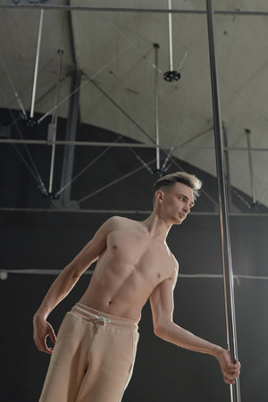 Vertical low angle view of bare to waist young Caucasian performer leaning on pole in dance studioの写真素材