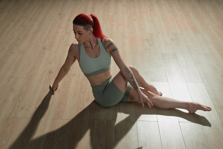 High angle view of tattooed Caucasian girl with red ponytail stretching hips sitting on studio floor...