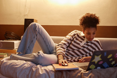 Full length portrait of Black teenage girl lying on bed at home and using laptop at nightの写真素材