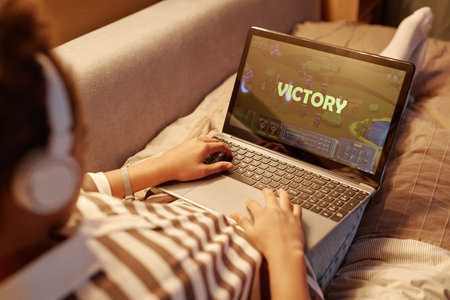Close up of teenager playing online video games via laptop with victory screen while lying on bed at nightの写真素材