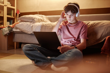 Full length portrait of young teenage boy using laptop while sitting on floor at night with legs crossed and wearing wireless headphonesの写真素材