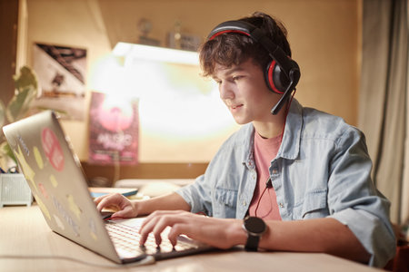 Side view portrait of teenage boy playing online video games with wireless headset at night and...