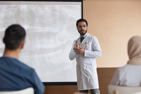 Bearded Muslim man doctor in glasses holding microphone giving speech at medical presentation with dental X-Ray on projection screen in backgroundの写真素材