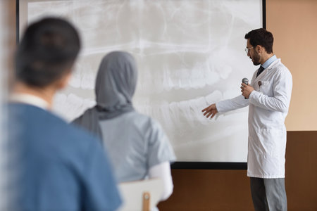 Medium full shot of Muslim man medic holding microphone showing dental X-Ray on projection screen to audience during medical conferenceの写真素材