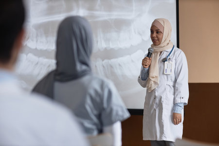 Young Islamic woman medical specialist in beige hijab talking into microphone addressing audience standing by projection screen with dental X-Ray during conferenceの写真素材
