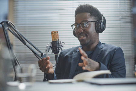 Medium close up shot of young Black man wearing headphones and eyeglasses speaking and adjusting microphone stand during live podcastの写真素材