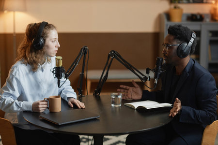Side view of Caucasian woman with cup talking to gesturing Black man in suit during podcast, both wearing headphones while sitting at round table in studioの写真素材