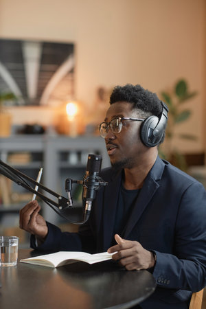 Vertical shot of African American male interviewer in headphones and glasses speaking into microphone gesturing with pen in hand sitting at desk in podcast studioの写真素材