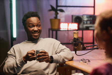 Medium shot of amused Black male influencer sitting in neon lit podcast studio with unrecognizable guest and holding coffee cupの写真素材