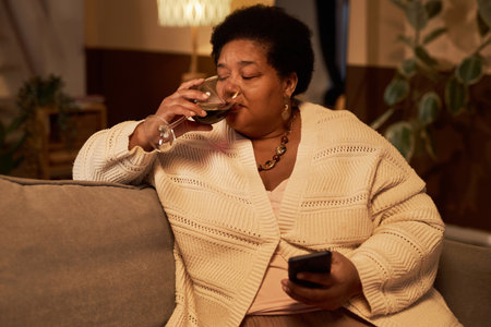 Portrait of African American senior woman using smartphone and drinking wine while enjoying relaxing evening at home, copy spaceの写真素材