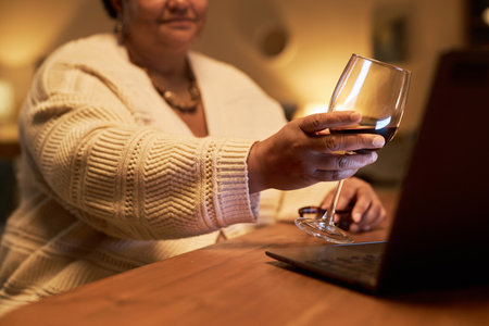 Close up of unrecognizable mature woman clinking wine glass to screen during online celebration with friend or partner, copy spaceの写真素材