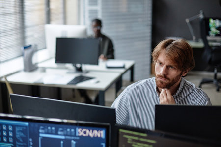 High angle portrait of bearded young man working with multiple computers in IT office setting copy...