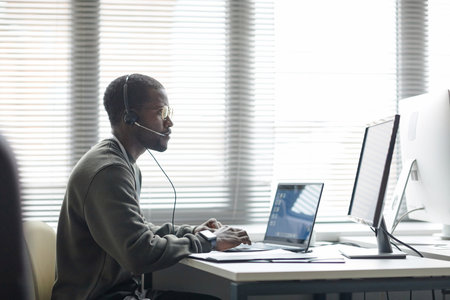 Side view portrait of Black young man wearing headset and using computers as client support operator...