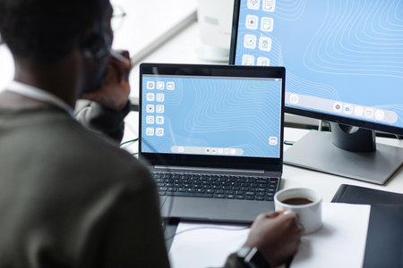 Over shoulder view of young Black man using laptop with blue desktop screen in minimal office...