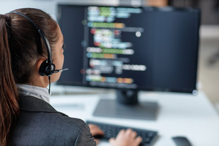 Over shoulder view of young woman wearing headset and using computer with code lines on screen in IT...