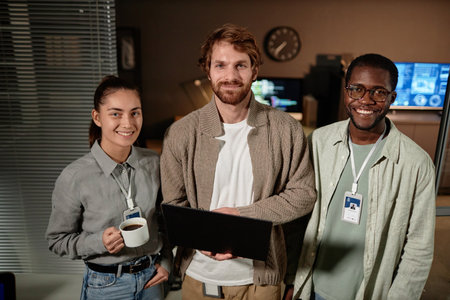 Waist up portrait of three young people holding laptop and looking at camera while standing in IT...