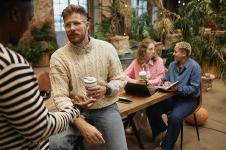 Portrait of young bearded businessman wearing casual sweater in office and chatting with colleague...