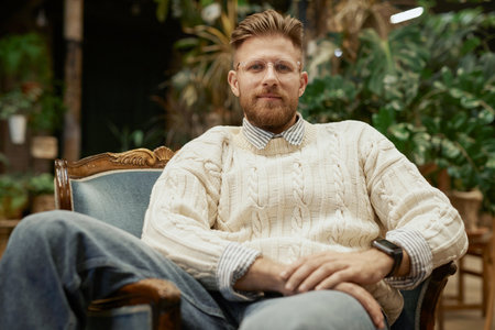 Portrait of bearded young man wearing glasses and casual sweater lounging in vintage chair and looking at camera with confidenceの写真素材
