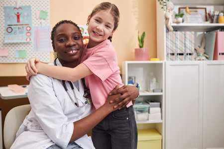 Portrait of Black young woman as doctor embracing little girl looking at camera in clinic officeの写真素材