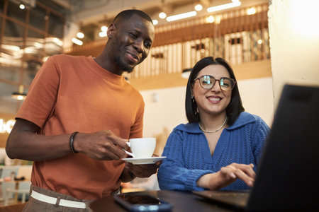 Waist up portrait of two smiling young people looking at laptop screen in coffee shop Black man and Middle Eastern womanの写真素材