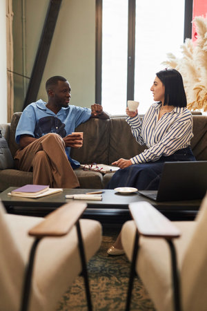 Vertical side view portrait of two diverse business people chatting in office lounge sitting on...