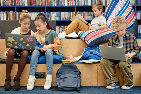 Full length view at group of young children enjoying break in modern library lounge in school and...