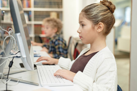 Side view portrait of serious young schoolgirl using computer in library during programming class...