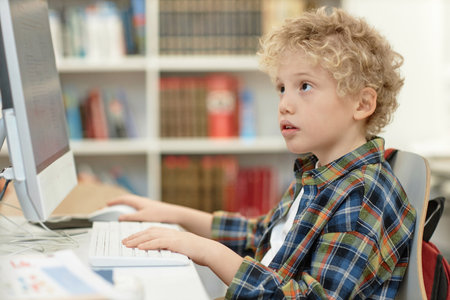 Side view portrait of cute curly haired boy looking at computer screen during online research or...