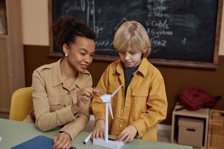 Portrait of young Black woman as smiling teacher showing wind turbine model to little boy and...